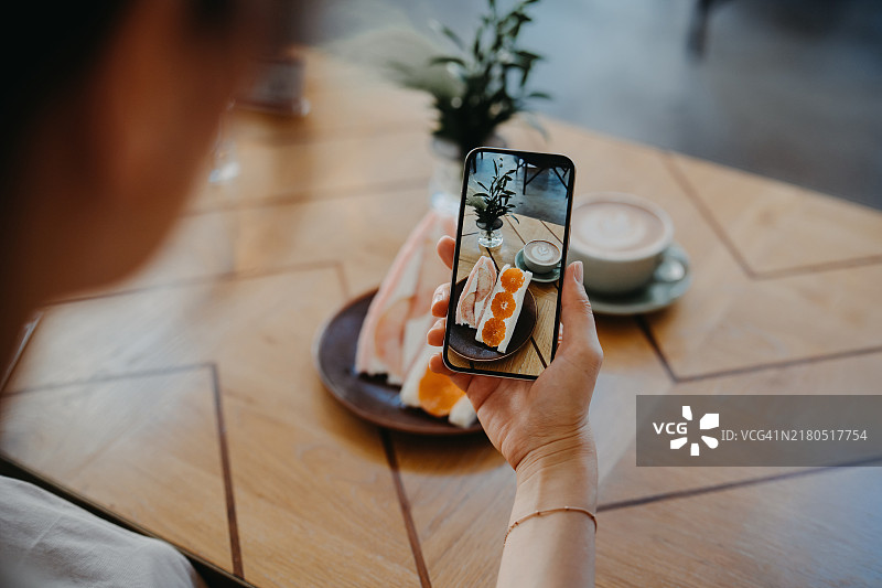 在咖啡店里，年轻亚洲女性用智能手机拍摄桌子上的水果三明治。数字餐饮。在社交媒体上分享美食照片。人物、食物、生活方式和技术图片素材