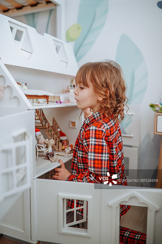 富有想象力的女孩穿着舒适的睡衣，在房间里玩着被彩色玩具包围的娃娃屋。图片素材