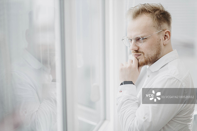 一位身穿白衬衫的年轻男商人若有所思地望着摩天大楼的窗外图片素材