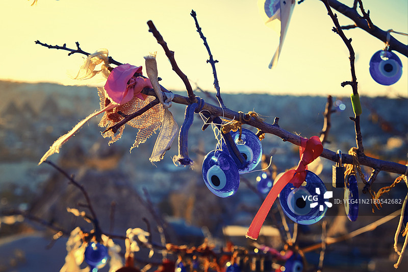 在日落的光芒中，树上装饰着纳扎尔和系着的丝带。纳扎尔是一种眼形护身符，据信可以防止邪恶之眼。土耳其卡帕多西亚内夫谢希尔省格雷梅图片素材