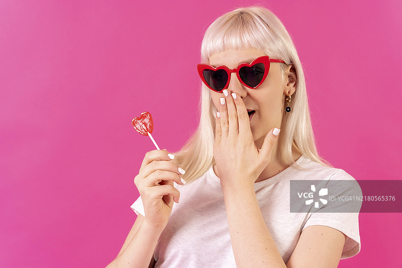 粉色背景工作室里的金发白人女孩，戴着心形眼镜，拿着棒棒糖图片素材