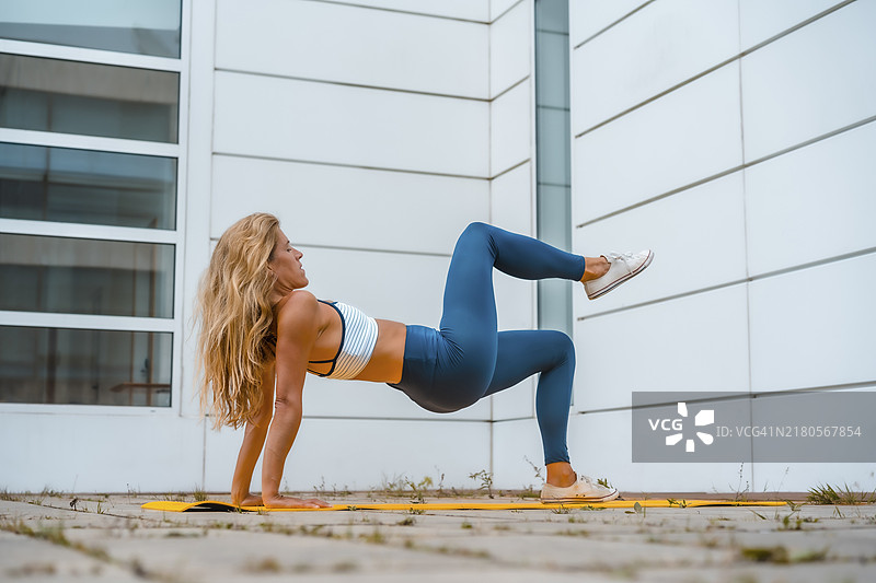 金发白人女孩在黄色垫子上锻炼，背景是白色墙壁，脚上穿着蓝色玛雅鞋，白色短款衬衫。面朝上弯曲右腿的健身场景图片素材