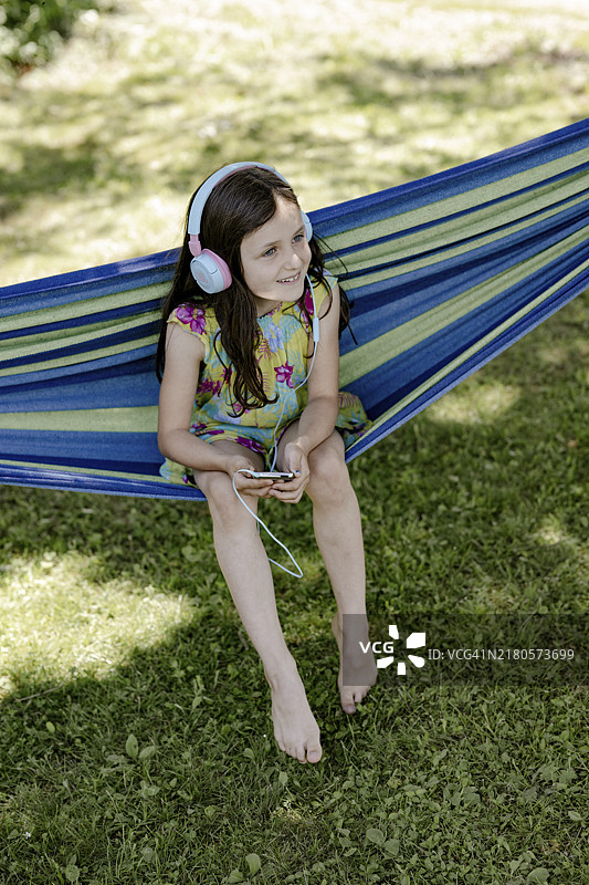 在阴凉处吊床上戴着耳机听音乐的8岁女孩图片素材