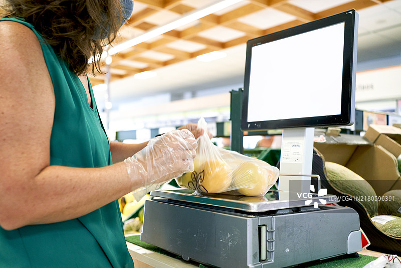 一位穿着休闲服的女性侧身站在购物中心，前景中正在称量一袋水果。图片素材