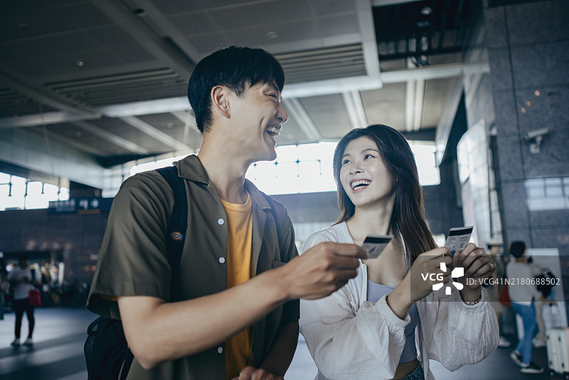 两位亚洲背包客查看当地高铁的出发时间，热切地讨论他们的旅行计划。他们站在售票柜台前，准备购买车票。图片素材