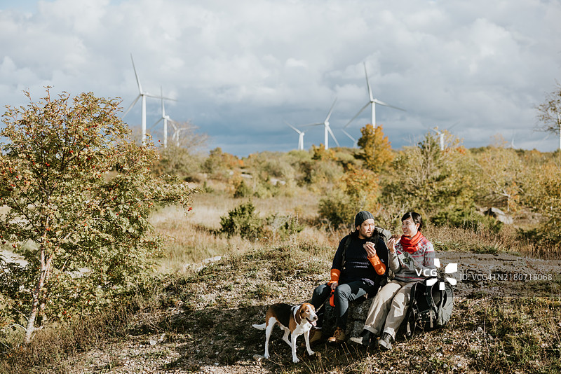 地平线上有风力涡轮机，两名徒步旅行者和他们的狗在旅途中享用三明治补充能量图片素材