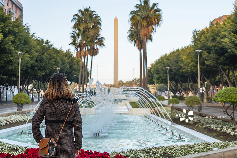在西班牙安达卢西亚阿尔梅里亚兰布拉的贝伦街，年轻女子看着喷泉和鲜花，旁边是地中海太阳海岸的棕榈树图片素材