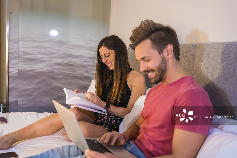 一对英俊的白人情侣在酒店床上休息，看电脑和书，夏季生活方式图片素材