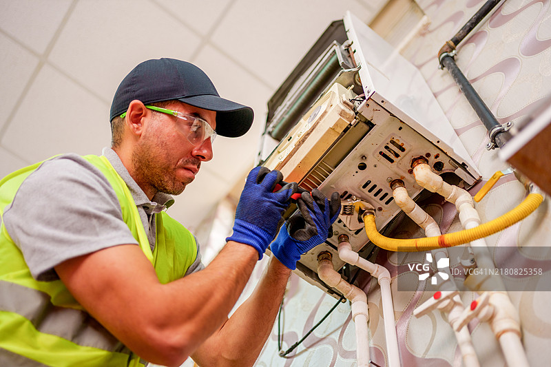 身穿红色背心和帽子的技术人员正在维修壁挂式锅炉的内部组件图片素材