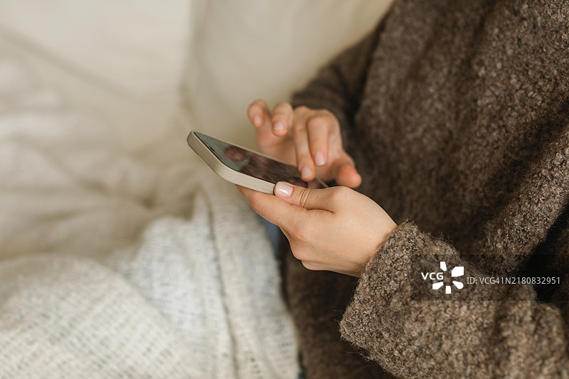 年轻女人穿针织套衫在沙发上，盖着毯子玩手机特写图片素材