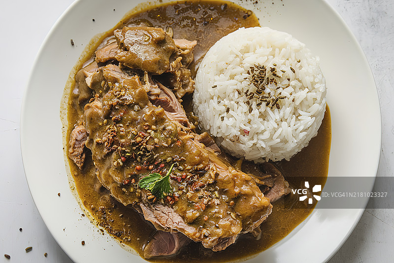 一只白色盘子里盛着肉汁炖猪肉，旁边配有一份熟白米饭。图片素材