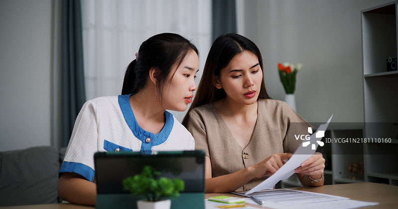 两位年轻的亚洲女性在客厅的桌旁使用平板电脑和文件查看家庭的财务交易图片素材