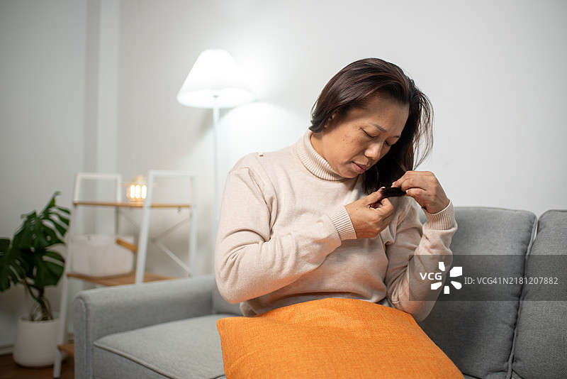 亚洲女性因脱发而感到沮丧，担忧的面容，双手抓着干枯、凌乱、毛躁的发尾，在家镜子前查看自己，健康与美容概念图片素材