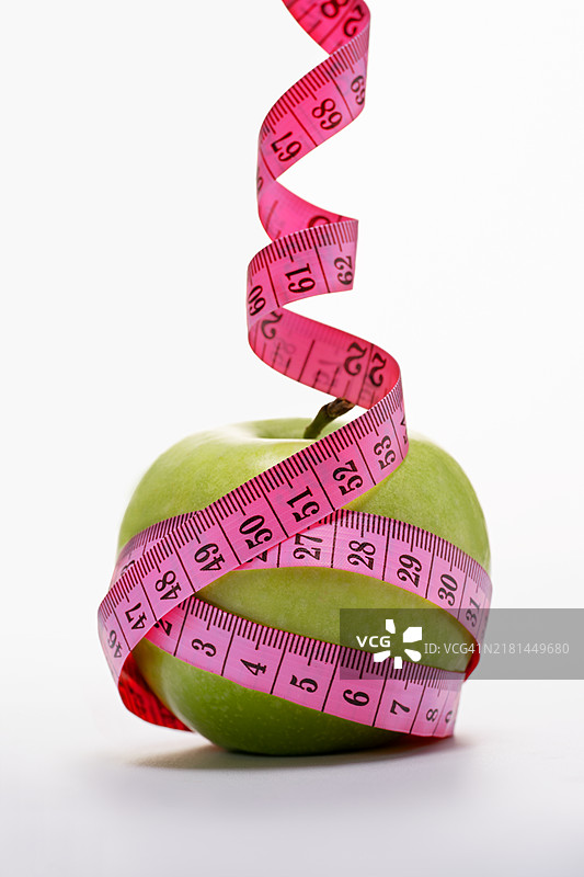 缠绕在青苹果上的粉色卷尺，白色背景。健康饮食理念，运动俱乐部。图片素材