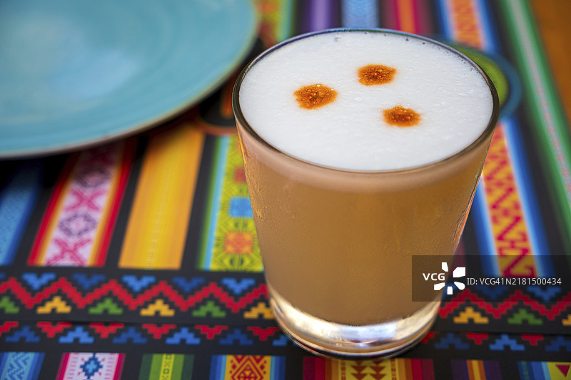 秘鲁皮斯科酸味鸡尾酒，由皮斯科（秘鲁葡萄烧酒）、酸橙汁、糖浆和蛋白制成，盛在带有传统图案的餐垫上的玻璃杯中图片素材