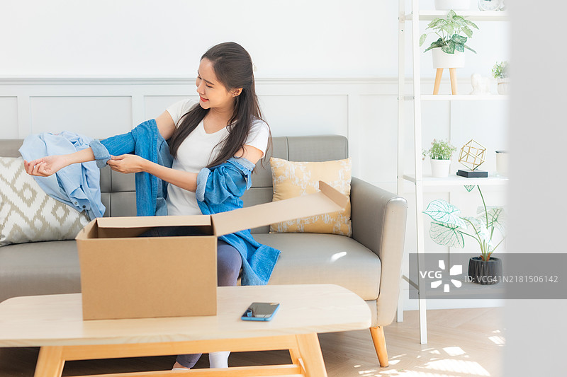 年轻的亚洲女性打开从网上商店收到的衣服。接收、打开并尝试在线购买的概念。图片素材