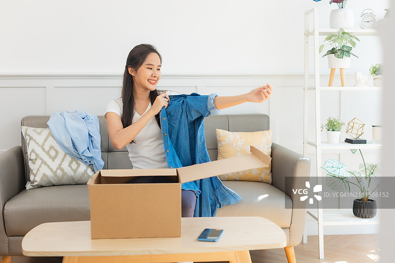 年轻的亚洲女性打开从网上商店收到的衣服。接收、打开并尝试在线购买的概念。图片素材