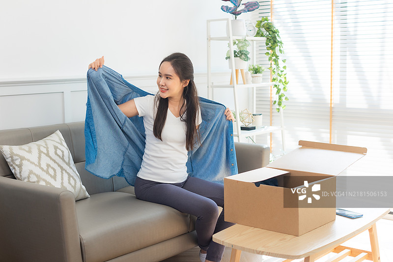 年轻的亚洲女性打开从网上商店收到的衣服。接收、打开并尝试在线购买的概念。图片素材