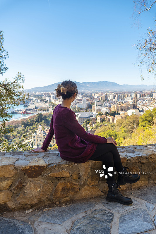 在西班牙马拉加吉布拉法罗城堡，年轻游客坐在春天里欣赏马拉加市和化身大教堂的景色图片素材