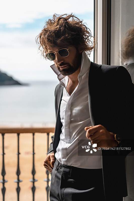 街头风格：豪华酒店卧室窗边，身穿黑色西装和白色衬衫的年轻白人男子，佩戴太阳镜，时尚大片图片素材