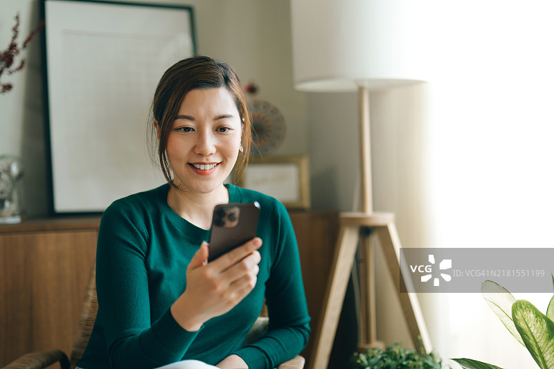 美丽的年轻亚洲女性在网上冲浪并使用智能手机进行在线购物。她坐在家中的扶手椅上使用手机。在线购物，电子银行。数字支付。移动电子商务。科技智能银行。图片素材
