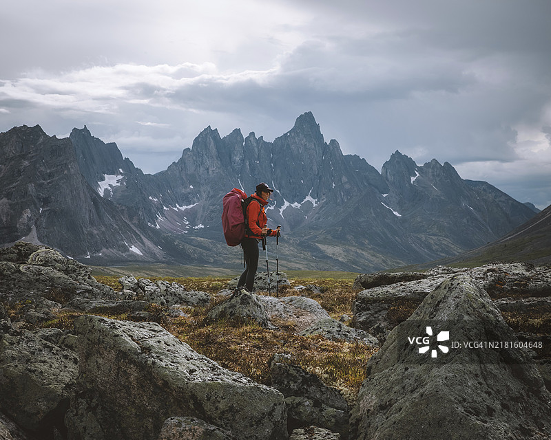 在多云天空下崎岖山地景观中，带着背包和登山杖的徒步旅行者，加拿大育空地区墓碑领地公园的妇女图片素材