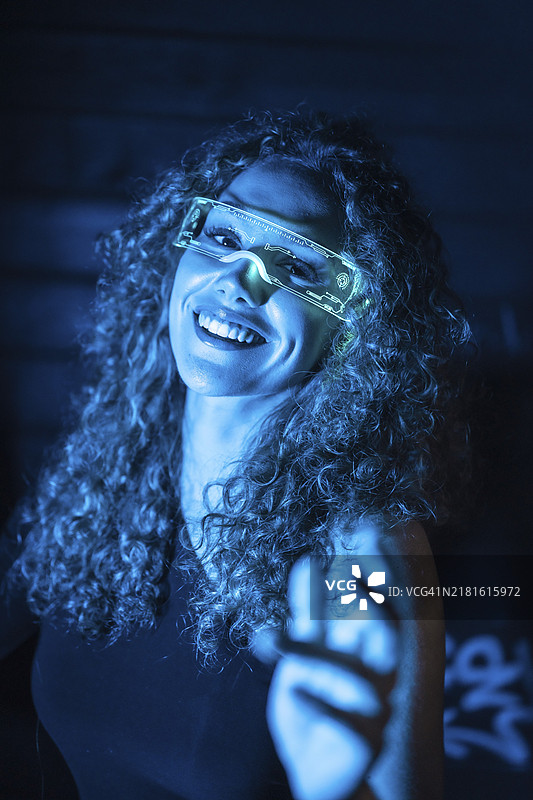 带有蓝色霓虹灯的未来主义快乐年轻女性戴着智能护目镜做手势的竖版特写肖像图片素材