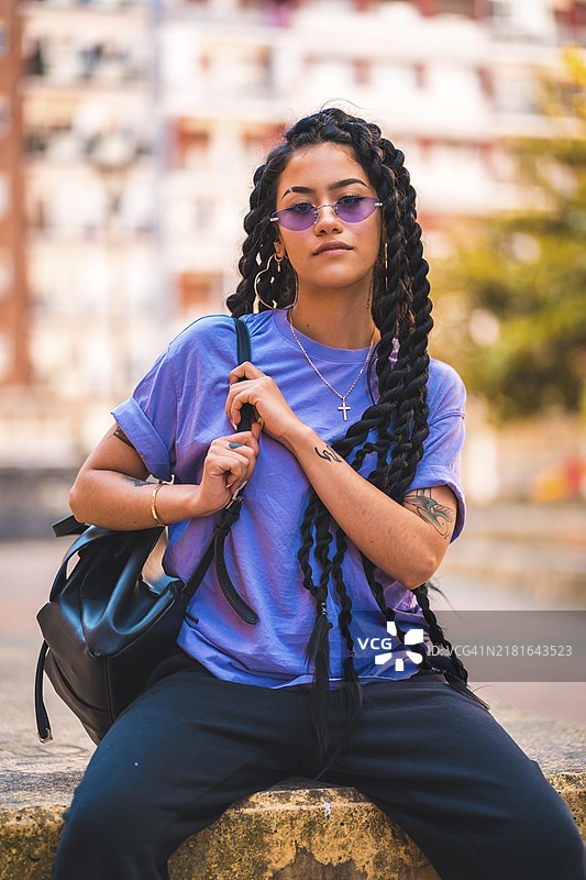 生活方式：年轻的深色皮肤女人，留着长辫子，戴着紫色眼镜，坐在公园的长椅上，肩上挎着背包图片素材