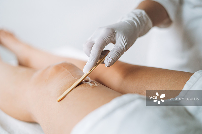 年轻美女专家为躺在美容院沙发上的女性腿部涂抹脱毛凝胶图片素材