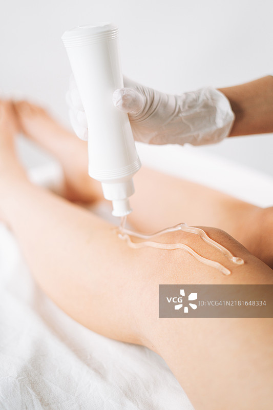 年轻美女专家为躺在美容院沙发上的女性腿部涂抹脱毛凝胶图片素材