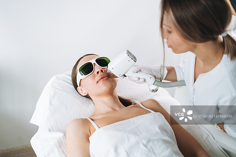 年轻女性戴着护目镜，躺在美容院的沙发上，用激光去除面部上唇的毛发。图片素材
