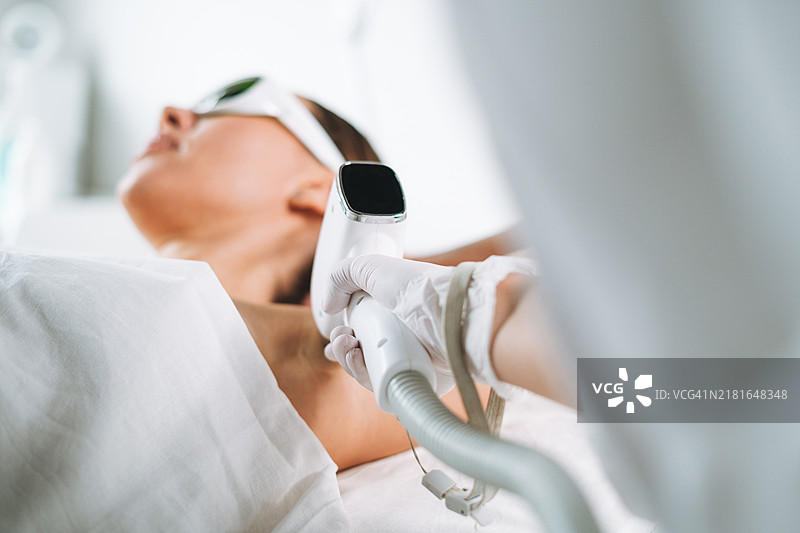 年轻女性戴着护目镜，躺在美容院的沙发上用激光去除腋毛。图片素材