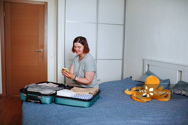 中年丰满女性手持智能手机，床上放着打包好的行李箱，正在寻找住宿并准备旅行。图片素材