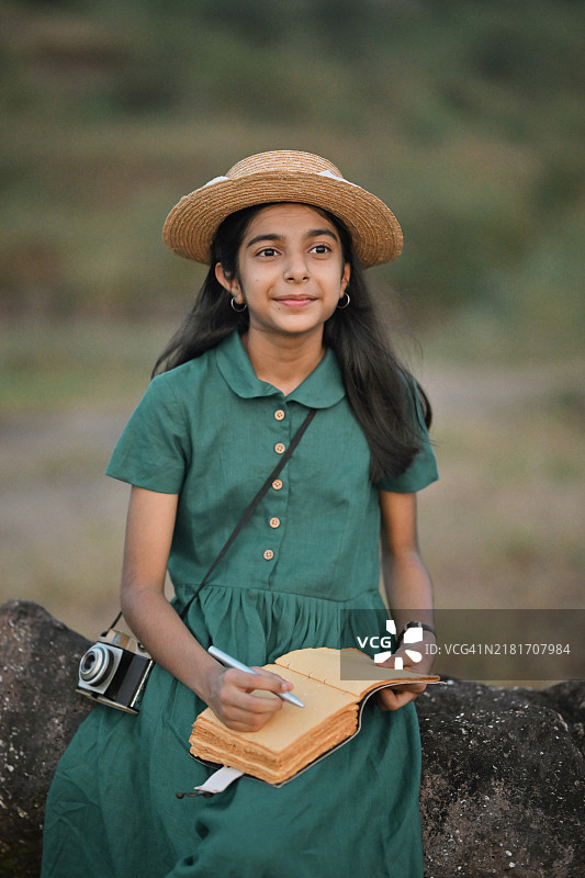穿着经典绿色亚麻裙和草帽的快乐女孩，手里拿着一本打开的书和一支笔，置身于大自然中。图片素材