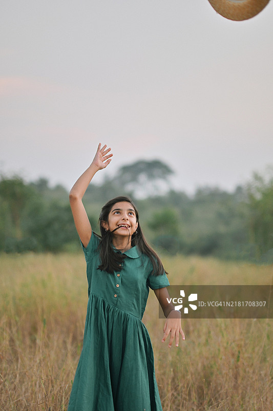 穿着经典绿色亚麻裙的小女孩在大自然中将草帽抛向空中图片素材