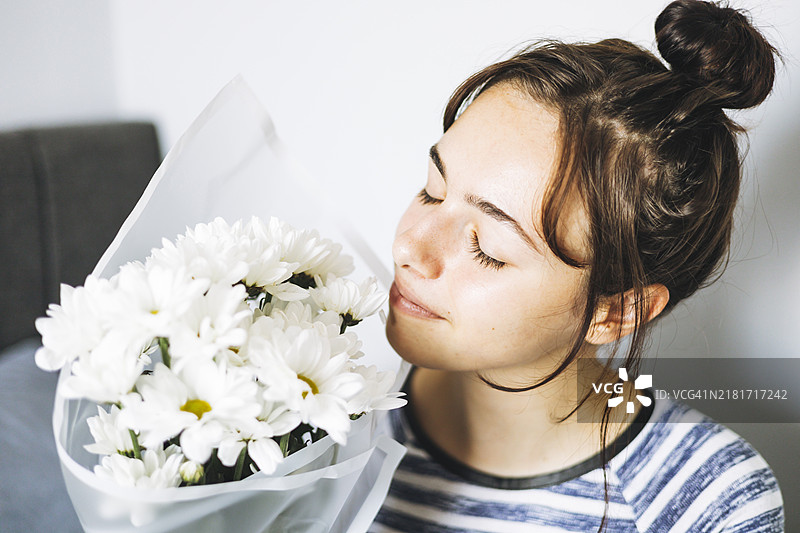 年轻女性在温馨的室内环境中微笑着，手中捧着一束白色雏菊，阳光明媚。图片素材