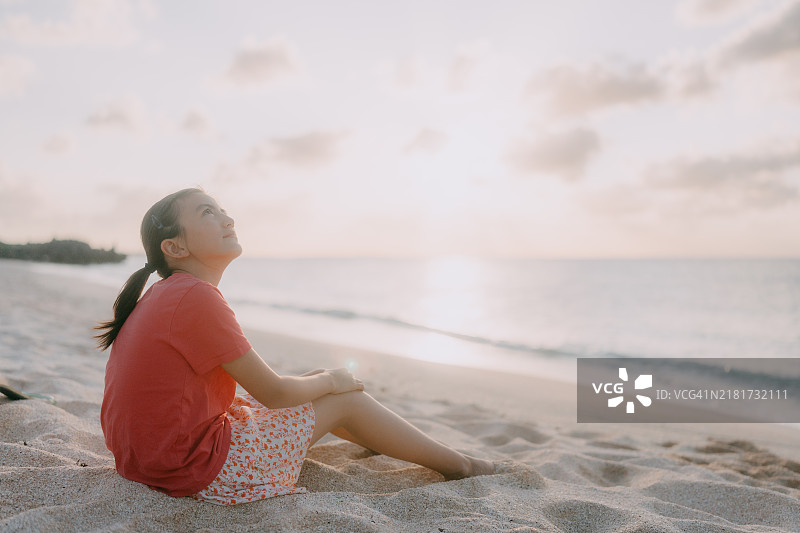 年轻女孩坐在海滩上，仰望着夕阳下的天空图片素材