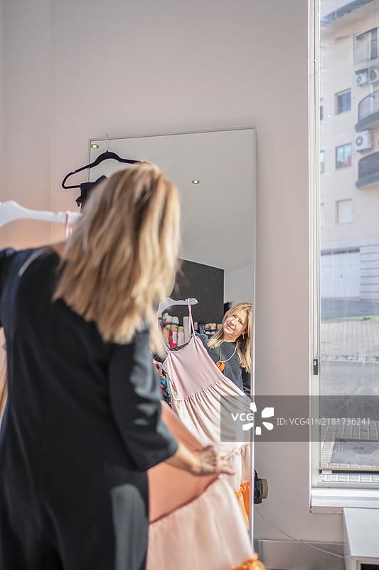 一位时装设计师在明亮的工作室里，手持一件粉色和橙色的裙子，站在镜子前。垂直拍摄，突显创意工作空间和服装设计过程。图片素材