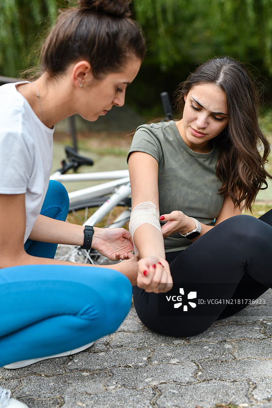 从自行车上摔下来后不适坐在地上的女骑行者，女朋友正在用绷带包扎她的手图片素材