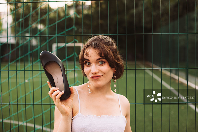 年轻女性在足球场旁的围栏边拿着高跟鞋的肖像图片素材
