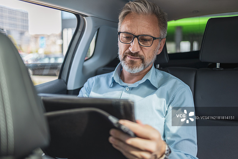 中年男商人通勤中在出租车后座用平板电脑远程办公。他戴着眼镜，穿着蓝色衬衫，看起来很专注于工作图片素材
