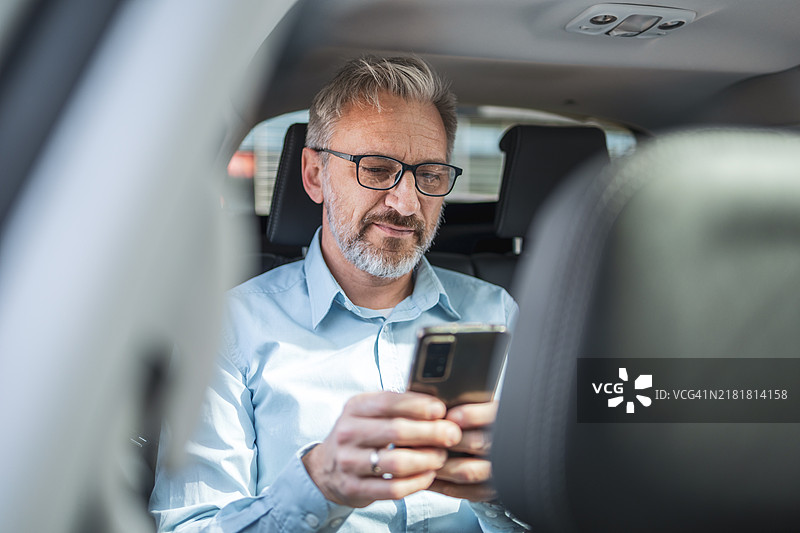 成熟的商人坐在出租车后座远程工作，使用智能手机。该男子穿着浅蓝色衬衫和眼镜，在通勤时专注于工作图片素材