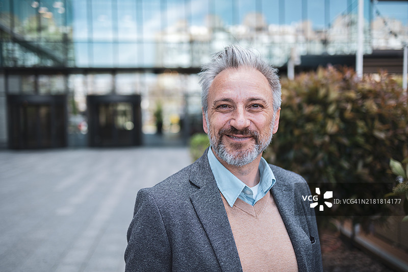 自信的成熟商人站在现代办公楼前，面带微笑，传达出专业和成功的感觉，适合商业相关概念和企业视觉效果图片素材