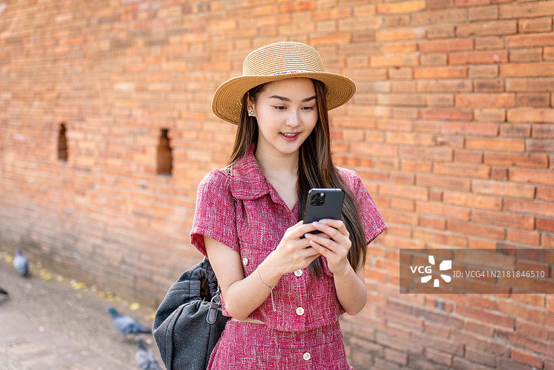 快乐的亚洲女游客戴着帽子在清迈塔佩门附近的历史砖墙边使用手机，周围环绕着鸽子图片素材
