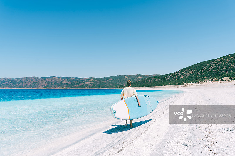 年轻男子手持蓝色划水板在蓝色海洋旁散步。图片素材