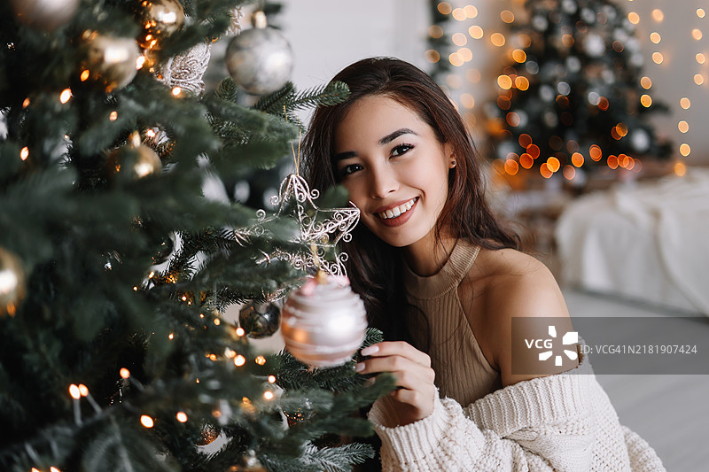 一位亚洲女性在温馨的客厅里用气球和玩具装饰圣诞树，为冬季的圣诞节做准备。图片素材
