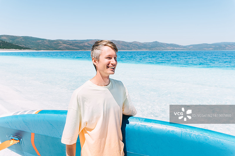 年轻男子手持蓝色划水板在蓝色海洋旁散步。图片素材