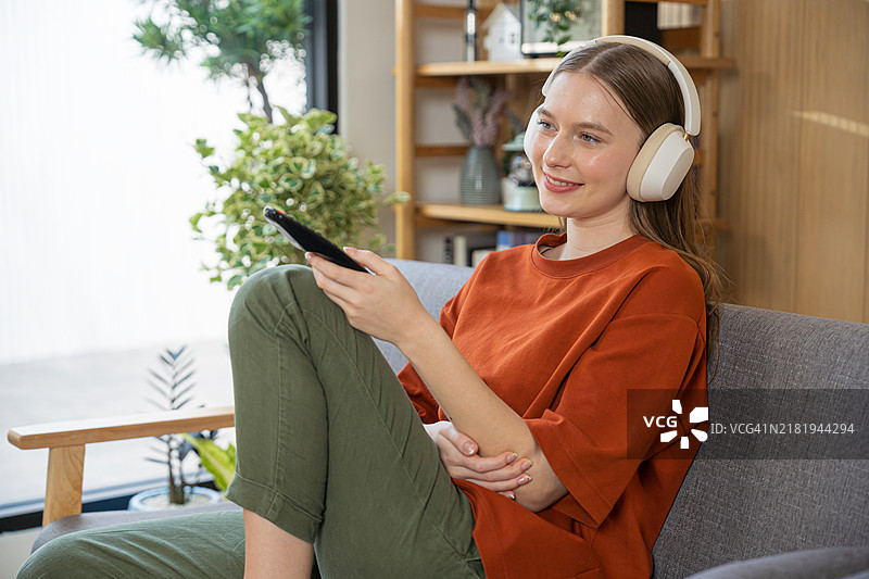 美丽微笑的女性戴着耳机，正在看电视并握着遥控器。图片素材