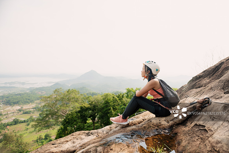 亚洲女性攀岩者坐在悬崖边缘休息，欣赏山景。图片素材