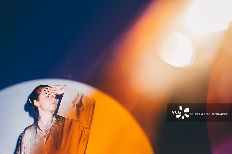 身穿丝绸衬衫的女子，手遮住眼睛，沐浴在戏剧性的彩色光影投射中，橙色、紫色和白色的混合色调营造出艺术感和电影感。图片素材